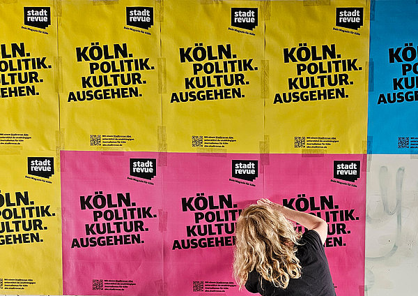 Foto: Elke Schwirtheim / Stadtrevue Eine Frau beklebt eine Wand mit bunten Plakaten. Darauf steht: "Köln, Politik, Kultur, Ausgehen — Stadtrevue, dein Magazin für Köln"